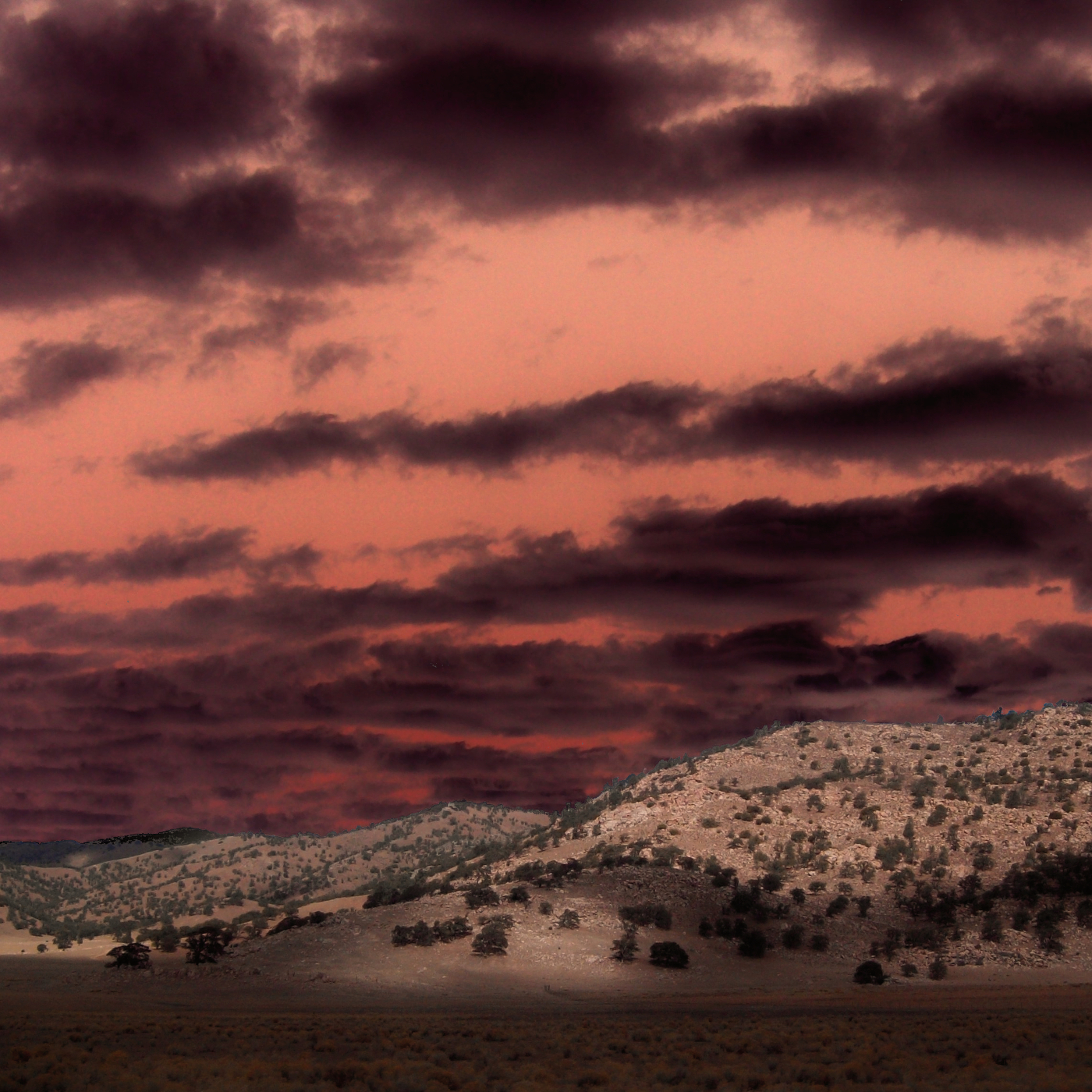 Distant landscape view of mountains from a desert climate. The sky above is tinted red and has many black clouds.
